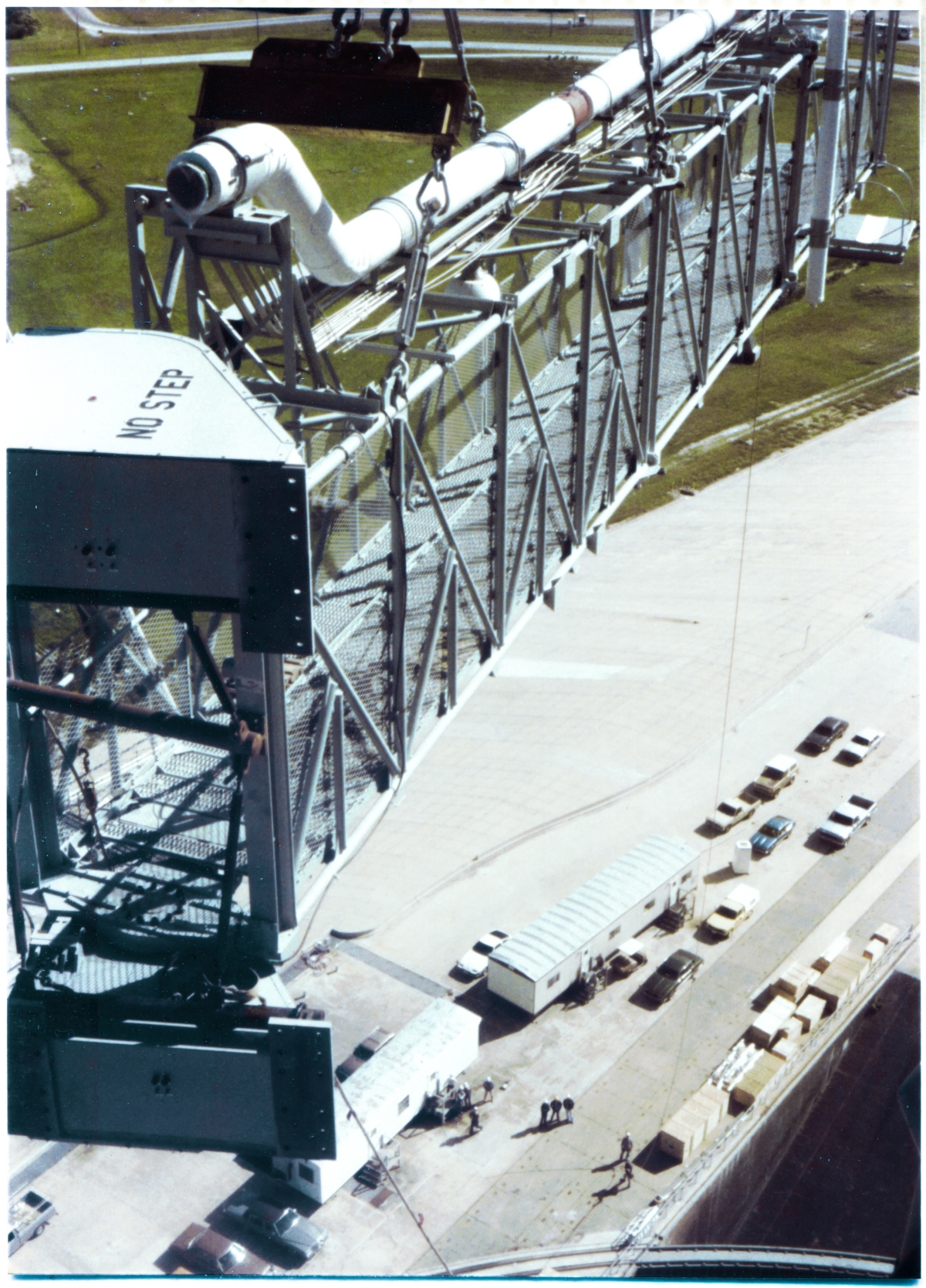 Image 110. At Space Shuttle Launch Complex 39-B, Kennedy Space Center, Florida, the Gaseous Oxygen (GOX) Vent Arm is closing in on its destination near the top of the Fixed Service Structure, lifted by the Hammerhead Crane, and controlled in orientation by the Union Ironworker visible far below on the ground, holding the tag line which prevents the Arm from swinging around into any undesired orientation. You're looking down at things from Camera Platform 8 at elevation 285'-0” on the northeast corner of the FSS, and to your right and below you, just out of frame, ironworkers are already in place to make the connection, once the Arm comes into direct contact with the Strongback which it will be bolted to. Just prior to that, the ironworker holding the tag line will walk to the north, rotating the arm to an orientation where the Connection Plates that are visible on the Arm's Hinge Boxes will line up flush with the drilled surfaces of the Strongback Columns it will be attached to, and at that point the ironworkers on the FSS will insert bolts through the holes in the Strongback and the Connection Plates you see in this image, and then torque them down, permanently affixing the Arm to the tower. Photo by James MacLaren.